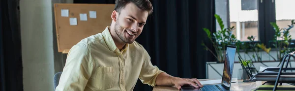 Позитивный бизнесмен в рубашке с помощью ноутбука и глядя на камеру в офисе, баннер — стоковое фото