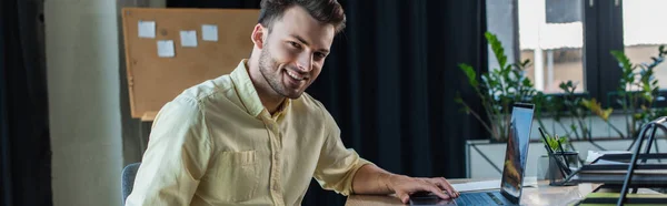 Positive businessman in shirt using laptop and looking at camera in office, banner — Fotografia de Stock