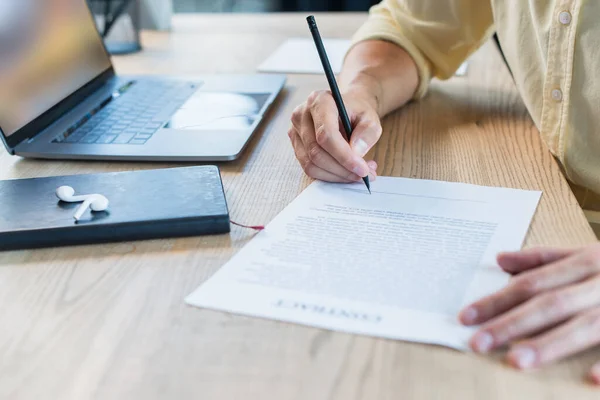 Обрезанный взгляд бизнесмена, пишущего на размытом контракте возле ноутбука и наушников на столе — стоковое фото