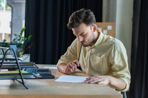 Pensive businessman в сорочці дивиться на документ біля ноутбука і документи в офісі — стокове фото