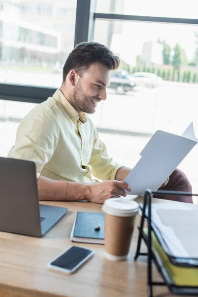 Вид сбоку улыбающегося бизнесмена, смотрящего на бумажную папку рядом с устройствами и кофе в офисе — стоковое фото