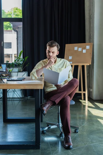 Задумчивый бизнесмен смотрит на бумажную папку рядом с ноутбуком на столе в офисе — стоковое фото