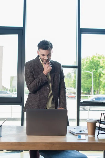 Улыбающийся бизнесмен в куртке смотрит на ноутбук рядом с ноутбуком и бумажной чашкой в офисе — стоковое фото