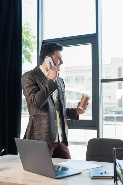 Side view of businessman talking on smartphone and holding paper cup near window in office — Fotografia de Stock