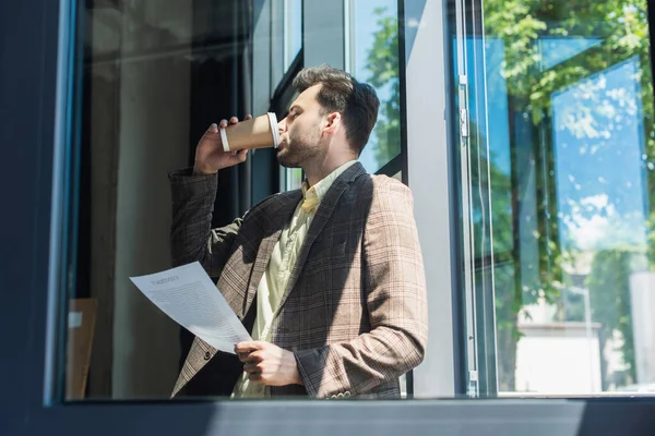 Side view of businessman holding document and drinking coffee to go in office - foto de stock