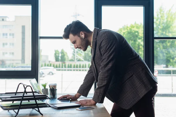 Side view of businessman in earphone using laptop near documents in office — Fotografia de Stock