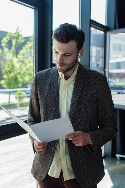 Young businessman in jacket using earphone and holding paper folder in office — Foto stock