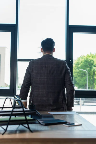 Обратный вид бизнесмена в куртке, стоящего рядом с устройствами и бумагами на рабочем столе в офисе — стоковое фото