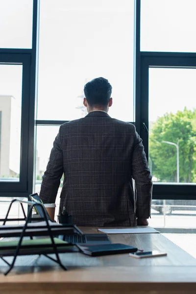 Back view of businessman in jacket standing near devices and papers on working table in office — Fotografia de Stock