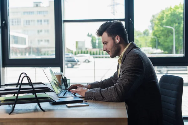 Side view of businessman in earphone using laptop near paperwork in office — Stockfoto