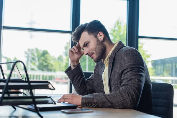 Side view of businessman in earphone using laptop near blurred papers in office — Photo de stock