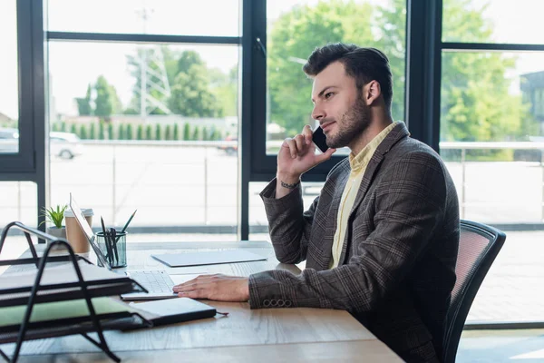 Side view of businessman talking on smartphone near laptop and papers on table in office - foto de stock