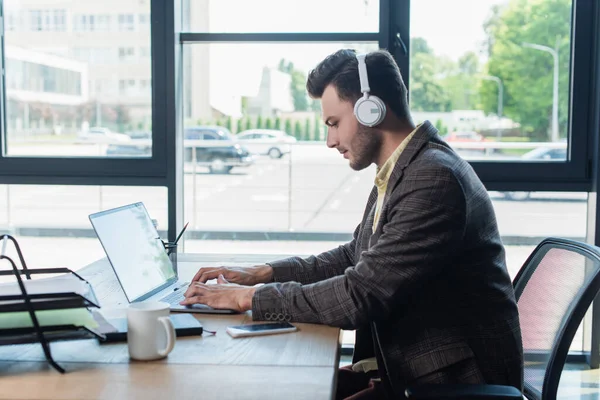 Side view of young businessman in headphones using laptop near paperwork and cup in office — Photo de stock