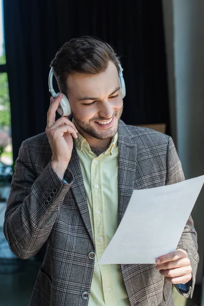 Позитивный бизнесмен слушает музыку в наушниках и держит документ в офисе — стоковое фото