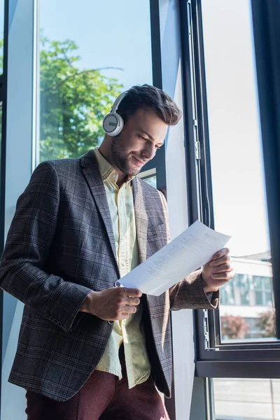 Young businessman in headphones looking at paper near window in office — Stock Photo