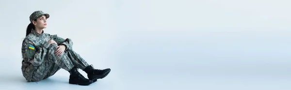 Servicewoman in camouflage uniform with ukrainian flag sitting on grey background, banner - foto de stock