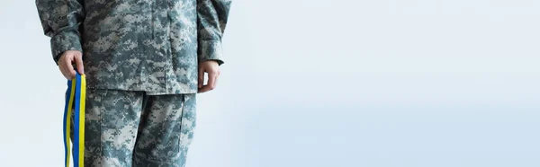 Cropped view of servicewoman in military uniform holding blue and yellow ribbon isolated on grey, banner — стоковое фото