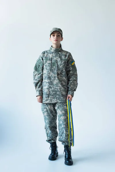 Full length of soldier holding blue and yellow ribbon and looking at camera on grey background - foto de stock