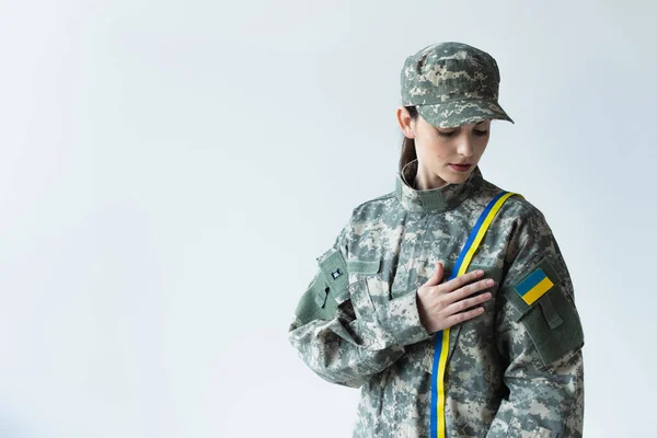 Young soldier in camouflage uniform with ukrainian flag touching blue and yellow ribbon isolated on grey — Stockfoto