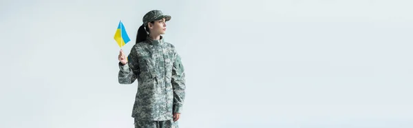 Soldier in uniform holding ukrainian flag and looking away isolated on grey, banner — Stock Photo