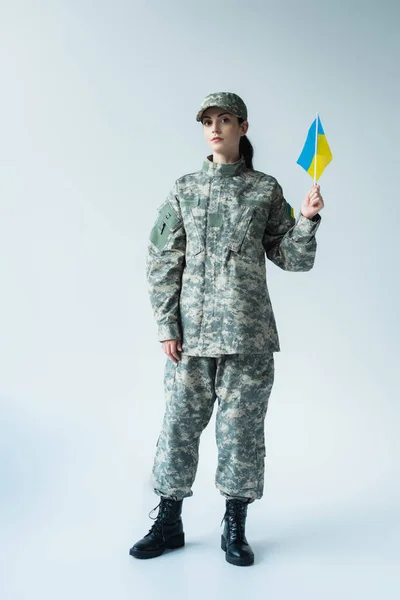 Young soldier in uniform and boots holding ukrainian flag on grey background — стокове фото