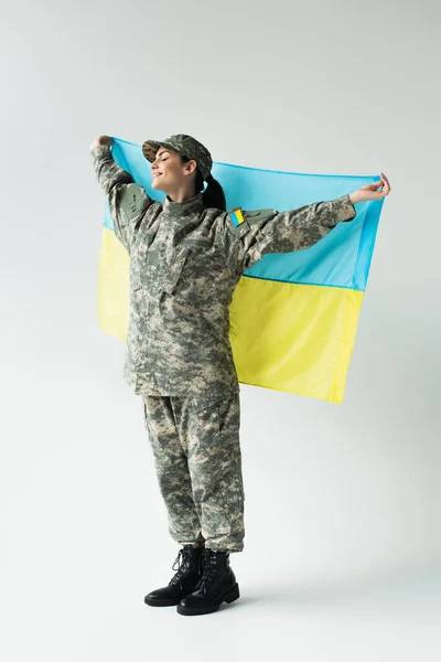 Side view of smiling soldier with closed eyes holding ukrainian flag on grey background — стоковое фото