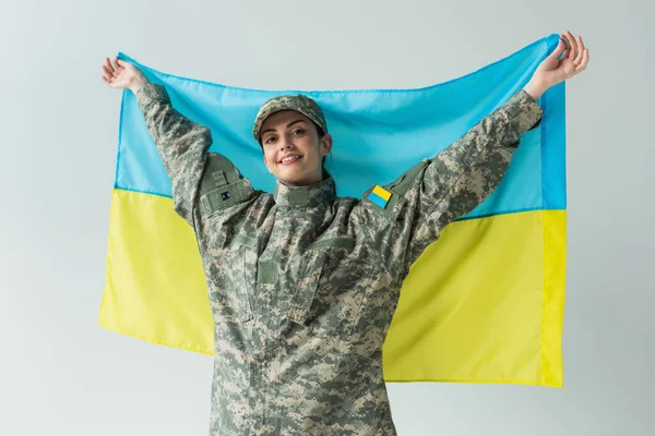 Smiling soldier in uniform holding ukrainian flag isolated on grey - foto de stock