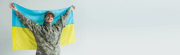 Happy servicewoman in camouflage uniform holding ukrainian flag isolated on grey, banner — Photo de stock