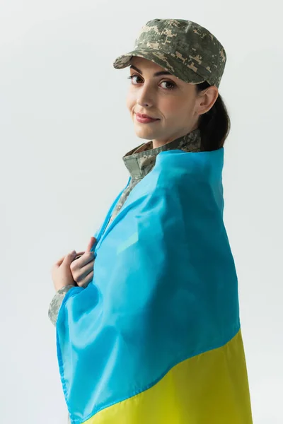 Young woman in military uniform covering with ukrainian flag isolated on grey — стоковое фото