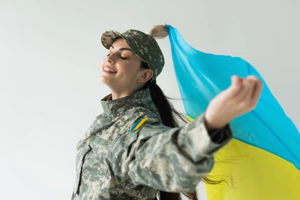 Cheerful soldier with closed eyes holding blurred ukrainian flag isolated on grey — стокове фото