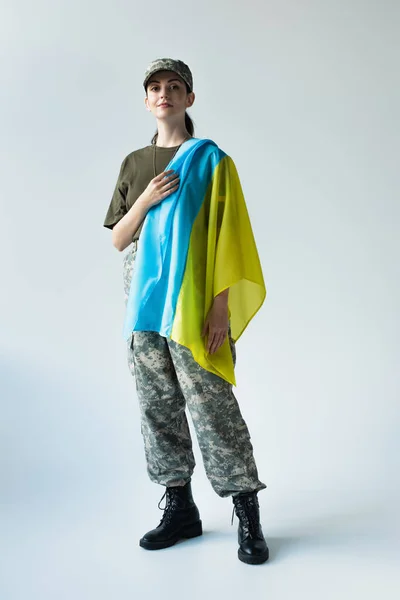Full length of young soldier with ukrainian flag looking at camera on grey background - foto de stock
