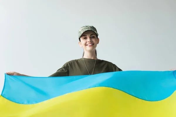 Позитивний солдат у шапці з україніанським прапором ізольований на сірому — стокове фото