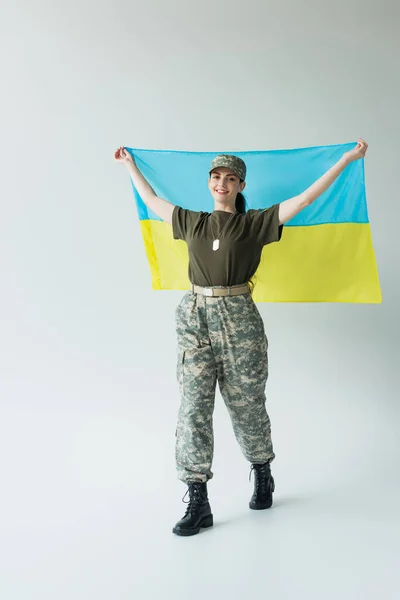 Smiling soldier in uniform and cap holding ukrainian flag while walking on grey background — Photo de stock