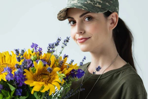 Portrait of smiling soldier with flowers looking at camera isolated on grey — Stock Photo