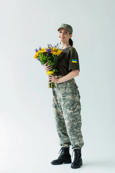 Positive soldier in uniform with ukrainian flag holding bouquet on grey background — Stockfoto