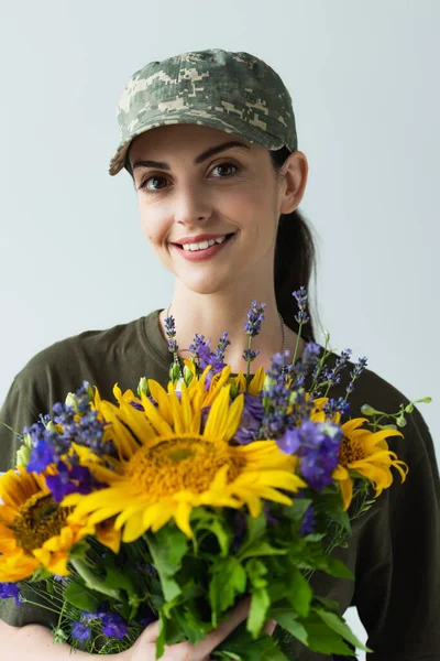 Портрет позитивної служниці в кепці, що тримає синьо-жовті квіти ізольовані на сірому — стокове фото