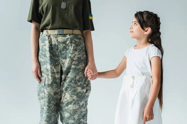 Smiling daughter holding hand of soldier with ukrainian flag on uniform isolated on grey — стоковое фото