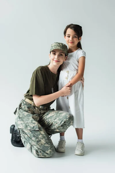 Mother in military uniform embracing smiling daughter on grey background — Photo de stock