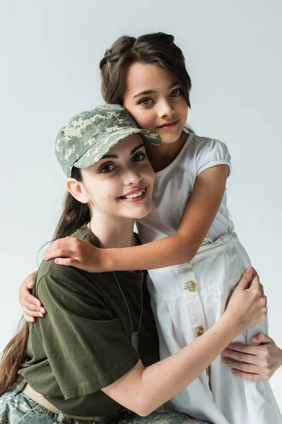 Smiling mother in military uniform looking at camera while hugging kid isolated on grey — Stock Photo