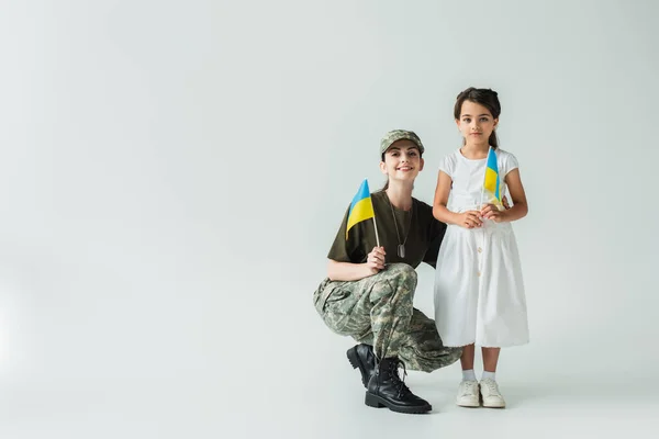 Soldier in uniform and daughter holding ukrainian flags on grey background — стоковое фото