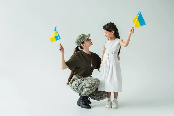 Side view of smiling parent in military uniform and child holding ukrainian flags on grey background — Fotografia de Stock