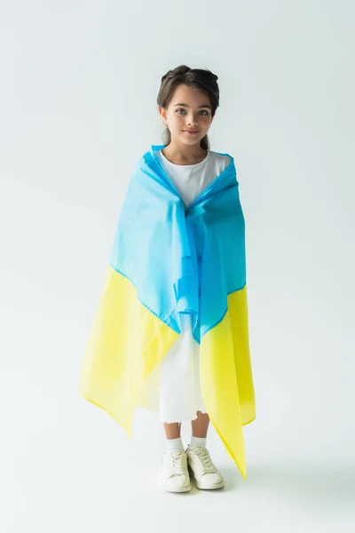 Smiling child covering with ukraining flag and standing on grey background - foto de stock