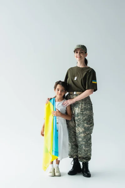 Full length of smiling woman in camouflage uniform hugging daughter with ukrainian flag on grey background — стокове фото