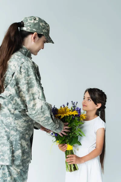 Kid giving blue and yellow bouquet to mother in camouflage uniform isolated on grey - foto de stock