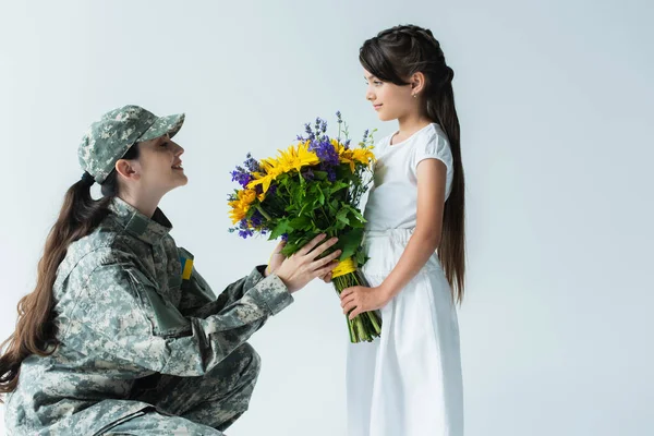 Вид збоку дитина дає синьо-жовті квіти мамі у військовій формі ізольовані на сірому — стокове фото