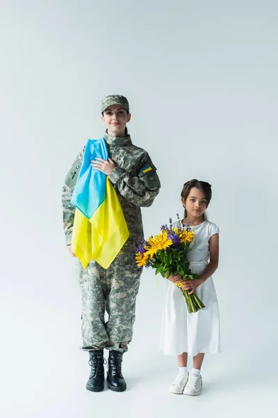 Солдат тримає український прапор біля дочки з букетом на сірому фоні. — стокове фото