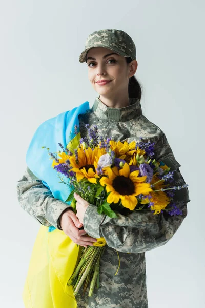 Усміхнений солдат з україніанським прапором, що тримає букет ізольований на сірому — стокове фото