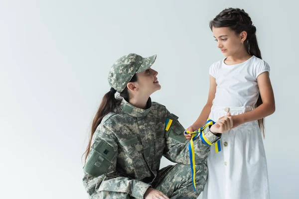 Kid holding blue and yellow ribbon and looking at mom in military uniform isolated on grey — стоковое фото