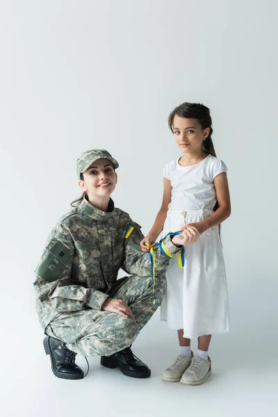 Smiling child holding blue and yellow ribbon near mom in camouflage uniform on grey background - foto de stock