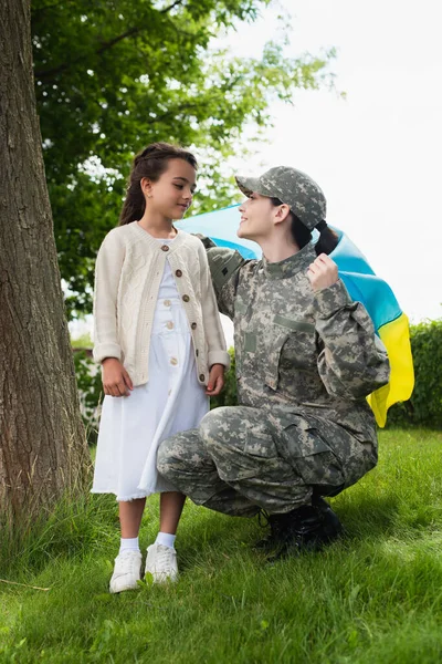 Smiling soldier holding ukrainian flag near daughter on grass outdoors — Foto stock